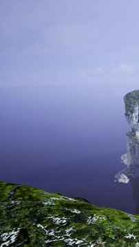 A tall cliff rises above the ocean with a rocky shoreline. The water below is calm. The sky is clear with few clouds. The scene captures the natural landscape and seascape.