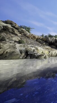 The scene shows a rocky shore with water reflecting the rocks and the sky. The greenery nearby adds to the natural setting during daylight, creating a simple landscape.
