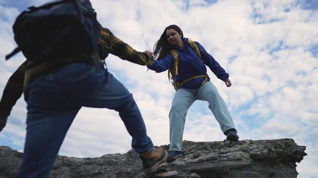Woman helping friend climb down rocky cliff during outdoor hiking adventure. Hikers with backpacks assist each other on rock formation. Friends help partner during mountain hike outdoors.