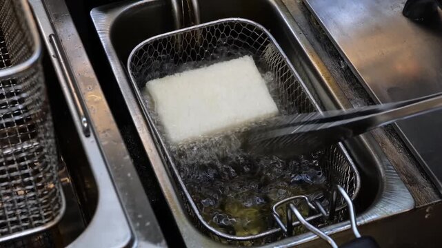 High-angle shot of a commercial-grade double tank deep fryer in a professional kitchen. The footage shows a metal wire basket containing a piece of food, submerged in bubbling, hot cooking oil. The st