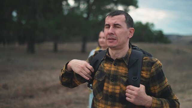 Male hiker wearing yellow plaid flannel shirt and black backpack shades eyes with hand while scanning distant horizon on wilderness trail. Outdoor explorer navigates forest path at dusk.