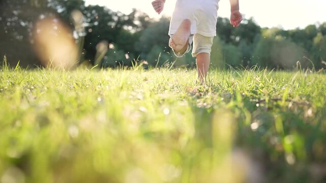 Child running barefoot on green grass in sunny park. Kid plays outdoors during golden hour. Barefoot toddler runs through meadow. Child enjoys nature in summer park. Kid playing on grass.
