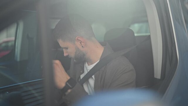 Man wearing seatbelt reaches with left hand toward steering wheel and turns head while seated inside car on street; focus alert.