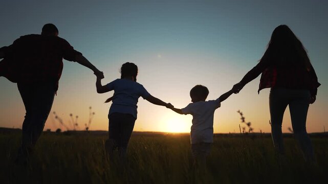 Family silhouette running together at sunset. Parents hold children hands while running in field. Silhouettes move across meadow. Family enjoys outdoor time during golden sunset light in nature.