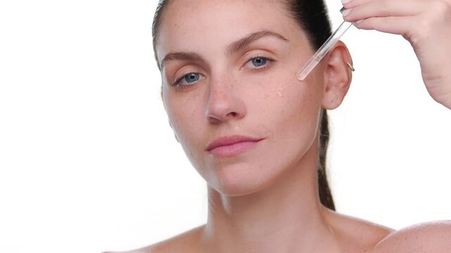 Zoomed in shot of Latina with light complexion and freckles applies hyaluronic acid face serum on her cheek with dropper against white bg | Face care hyaluronic acid commercial concept