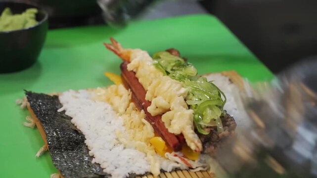 Close-up shot of a professional chef wearing black hygienic gloves preparing an authentic Japanese sushi roll (futomaki) or Korean kimbap. The chef is shown carefully layering crispy shrimp tempura, f