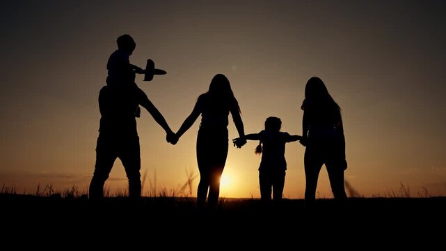 Beautiful family silhouette holding hands at golden sunset outdoors. Child sits on father's shoulders playing with toy airplane. Parents and children create lasting memories in nature's glow.