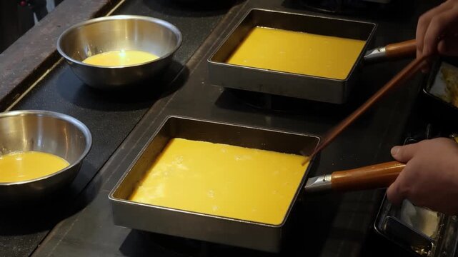 High-angle closeup of a chef's hands expertly cooking traditional Japanese rolled omelettes (tamagoyaki). The footage shows yellow beaten eggs being poured and rolled in specialized square pans on a p