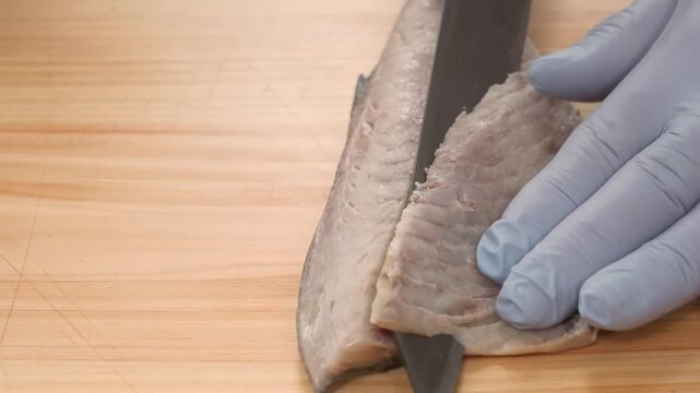 High-angle close-up of a chef in blue gloves delicately filleting a fresh saba (mackerel) on a clean wooden cutting board. The video demonstrates professional Japanese "Sanmai Oroshi" or three-piece f