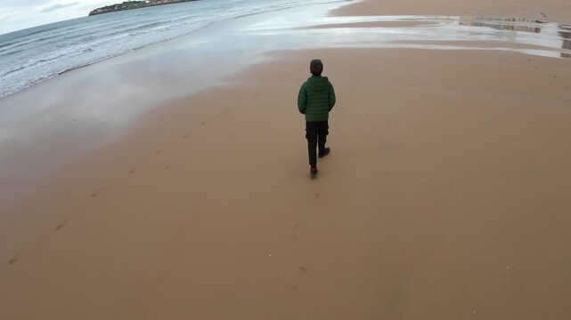Cinematic aerial style video of a teenage boy walking along a wide sandy beach near the ocean in northern Spain. Peaceful coastal atmosphere with travel and freedom concept.