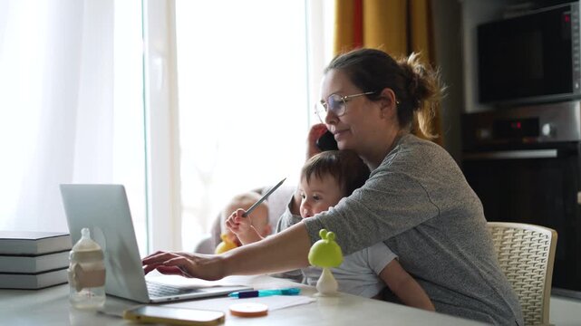 Mother multitasking with phone call while working on laptop and caring for two children at home. Woman balances remote work with childcare. Mom manages business tasks while kids need attention.