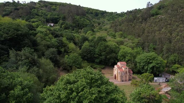 A cinematic drone zoom-in reveals San Miguel de Lillo in Oviedo, a UNESCO World Heritage site, rising above the trees and showcasing its Asturian pre-Romanesque beauty in a serene landscape.
