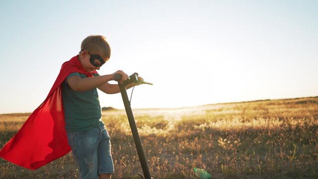 Superhero boy in red cape and mask stands with scooter in open field. Child pretends to be superhero at sunset. Kid smiles holding scooter handlebar outdoors. Superhero child plays in golden field.