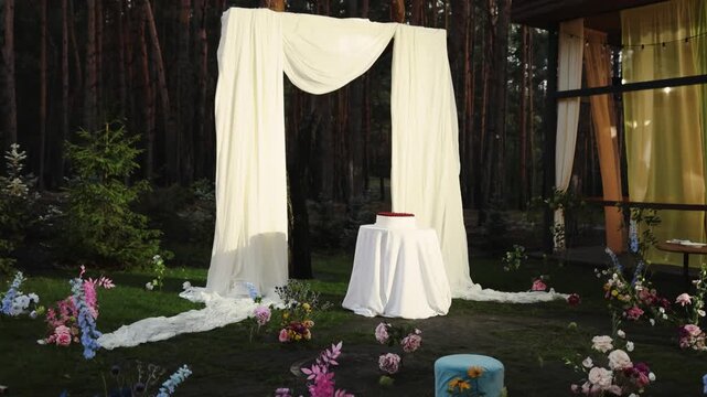 Wedding arch with white fabric drapery and elegant cake on table for outdoor marriage ceremony in pine forest. Beautiful rustic style venue decoration layout with fresh flowers bouquets on green grass