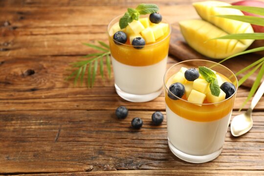 Delicious mango panna cotta with fruits and mint in glasses on wooden table, closeup. Space for text