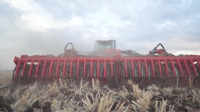 Red disc harrow works dry stubble field with tractor. Agricultural machinery cultivates soil for planting season. Farm equipment prepares land while dust clouds rise under dramatic cloudy sky.