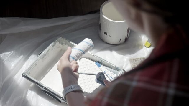 Woman fits paint roller cover onto handle beside tray, can, brush and white paint on protective sheet. Concept of painting preparation and renovation
