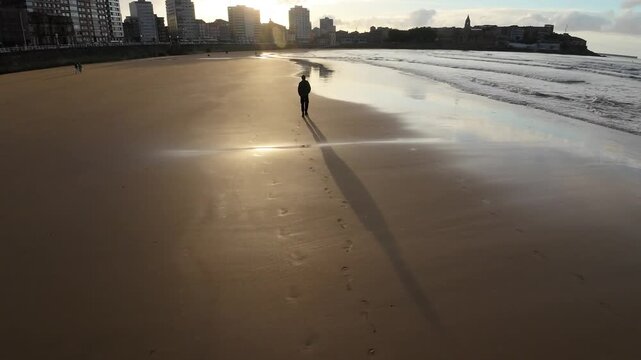 Aerial style view of a teenage boy walking along a sandy beach at sunset. Long shadow, ocean waves and peaceful coastal atmosphere in Gijon, Asturias, Spain.
