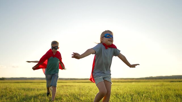 Children playing superhero running across green field wearing red cape and mask. Kids enjoy outdoor play pretending heroes. Active children run in meadow playing superhero game with capes.