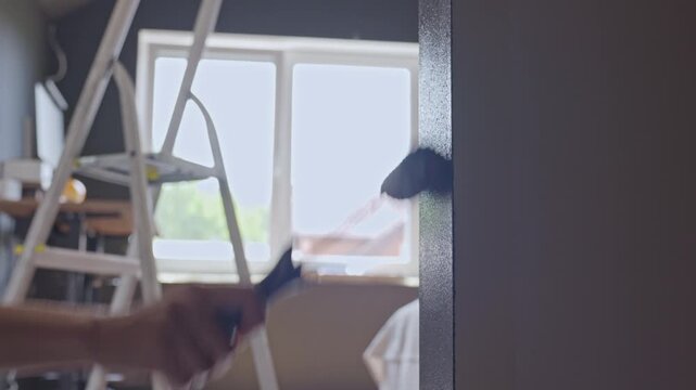 Person paints door frame edge with small roller near ladder and window. Concept of renovation, repair and painting work