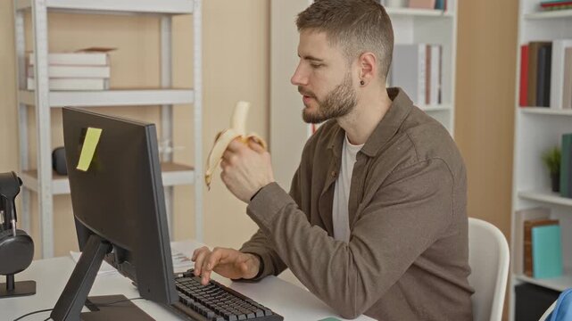 Man bearded wearing brown shirt typing at computer while holding peeled banana at desk in studio; concentration routine morning snack.