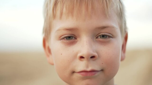 Blonde boy shows changing facial expressions while looking at camera. Young child displays different emotions outdoors. Kid with blonde hair shifts from serious to neutral expression face.