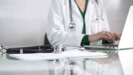 Fototapeta premium Female doctor using technology in healthcare, close up of hands typing on laptop with stethoscope, glasses, and clipboard on desk