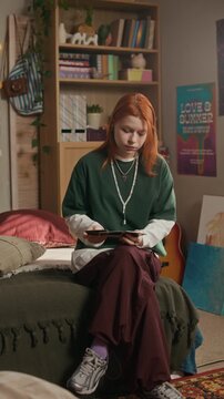 Vertical full portrait of Caucasian gen z girl with ginger hair wearing green long sleeve sitting on bed in stylish cozy bedroom with stack of polaroid photos in hands while looking at camera