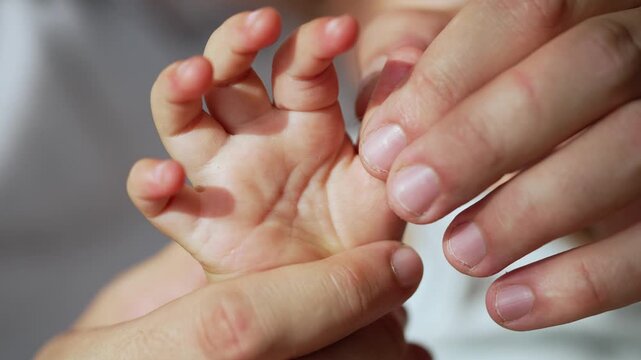 Baby hand moves tiny fingers close-up. Parent holds infant hand with gentle touch. Small palm and little finger detail visible. Baby and parent bond through hand contact. Tender care for newborn baby.