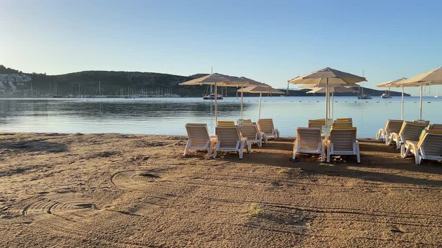 An empty sandy beach in Bitez, Bodrum, lined with neat rows of sun loungers and matching white umbrellas facing a calm, glassy blue bay beneath a bright morning sky.