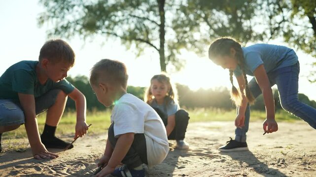 Children play and draw in sand with stick. Kids crouch on dirt path in park at sunset. Group of children play together outdoors. Kids explore nature in summer. Children draw on ground in golden light.
