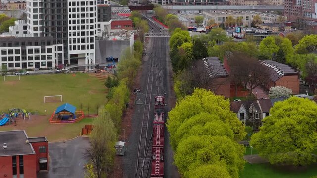 Urban freight train moving along railroad tracks through residential neighborhood in a canadian city with modern buildings, school buses and trees during overcast spring day