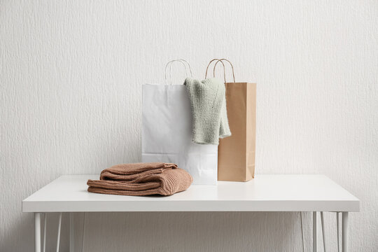 Paper shopping bags with stylish sweaters on table against white wall