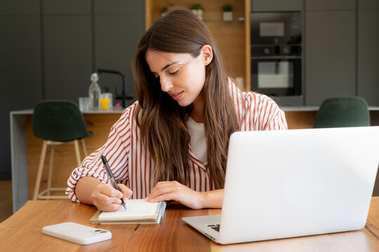 Young woman writing notes while working on laptop in modern kitchen home office