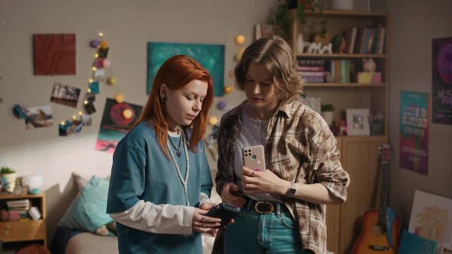 Medium shot of two young female friends taking selfie on smartphone and using portable instant printer to print photo while standing in cozy sunlit bedroom