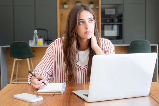 Young woman working from home on laptop taking notes and looking thoughtful in modern kitchen