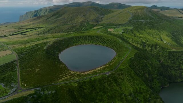 Aerial drone high-angle view of a perfectly circular volcanic crater lake surrounded by lush green caldera walls and hills on Flores Island, Azores.