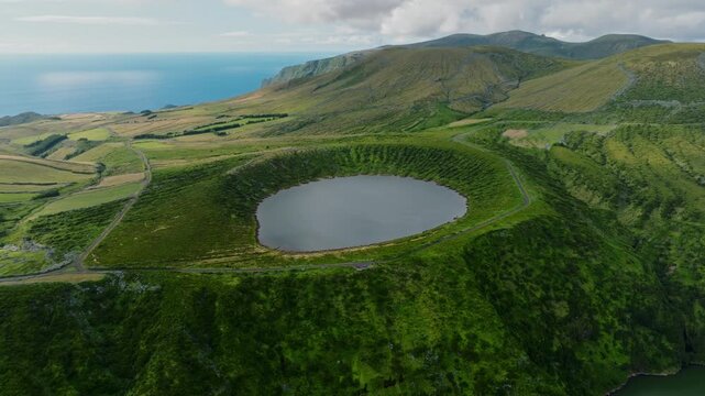 Title: Aerial drone wide view of a circular volcanic crater lake surrounded by lush green caldera slopes and a scenic road on Flores Island, Azores.