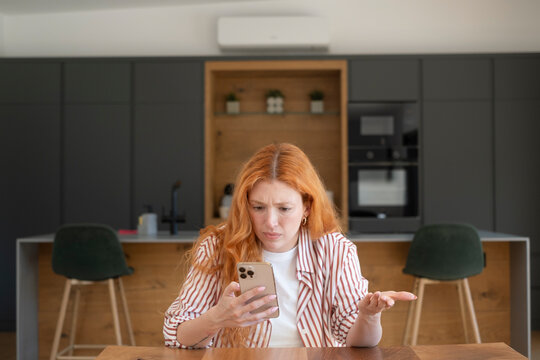 Frustrated redhead woman looking at smartphone in modern kitchen, confused by a message or error