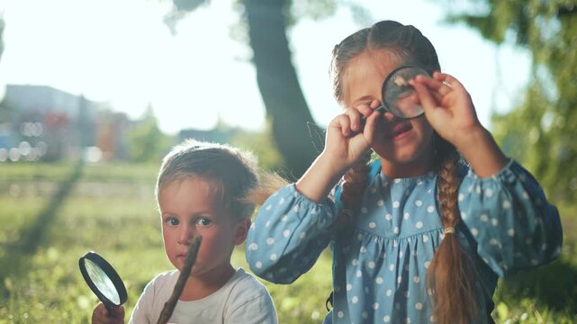 Children explore nature with magnifying glass in park. Girl looks through magnifying glass closely. Boy holds stick and magnifying glass on grass. Kids explore outdoors in summer sunlight together.