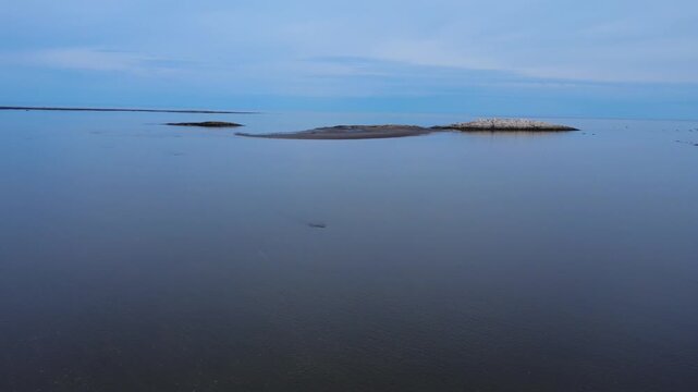 Drone footage of calm coastal waters at low tide revealing sandbars and rocky islets beneath a soft blue horizon in Mitis Bay, Quebec, Canada.