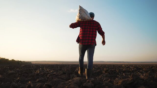 Farmer carrying sack walks across plowed field at sunset. Worker carries heavy bag on shoulder over tilled soil. Farmer in boots strides over farmland. Agricultural field stretches to clear horizon.