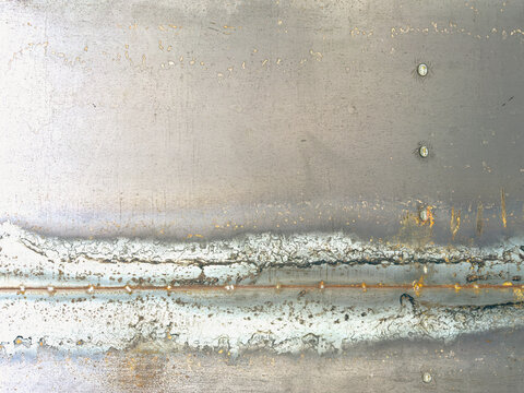 A textured industrial background showing a weathered silver metal sheet with detailed welding joints, burn marks, discoloration, rivets, and small spots of orange rust running along the seam