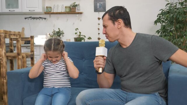 Father singing loudly into karaoke microphone on sofa. Daughter covers ear while dad sings. Man singing loud near child at home. Girl plugging ear on couch. Family karaoke fun in living room.