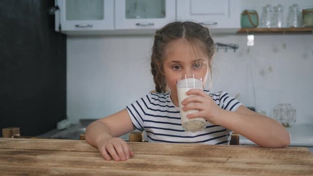 Girl drinking milk from glass in kitchen. Happy child sips milk at wooden table. Kid enjoys healthy drink and smiles. Little girl holds glass of milk. Child drinking beverage at home kitchen interior.