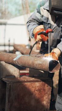 Professional welder in protective helmet joins large steel pipes outdoors with electric arc welding. Bright sparks and smoke illustrate industrial craftsmanship and durable metal construction process.