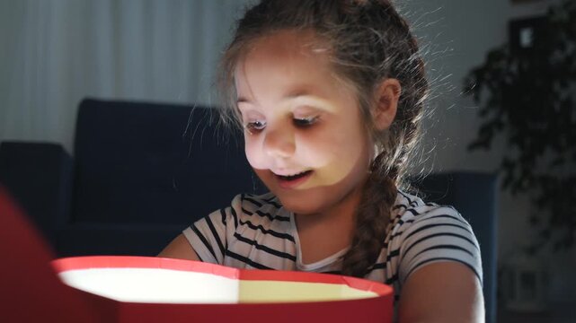 Happy child opens red heart box. Bright light from inside open box shines on face. Little girl looks at magic gift with surprise. Glowing light illuminates child. Wonder surrounds open gift box.