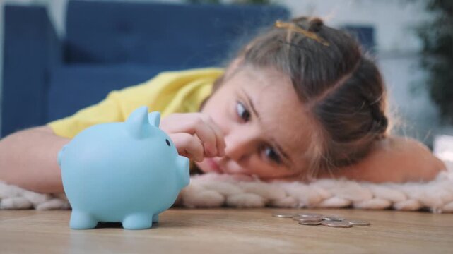 Child saving coins in piggy bank at home. Girl puts coin into blue money box on floor. Kid smiles while saving money. Child learns saving with piggy bank. Little girl collects coins for future saving.