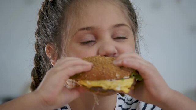 Hungry child eating delicious hearty burger. Little girl holding large hamburger with both hands. Kid taking huge bite of tasty fast food. Close up face shows child gladly enjoying eating burger.