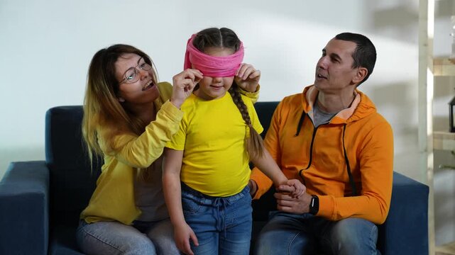 Family playing blindfold game on sofa. Mother ties pink scarf over child eyes. Parents enjoy fun play with daughter. Happy family bonding at home. Kid smiling with blindfold. Joyful time indoors.